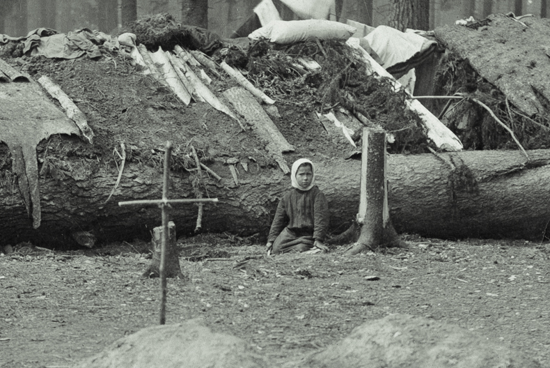 I förgrunden ett kors av pinnar nedstucket i jorden. I bakgrunden ett barn som sitter framför ett fällt träd som utgör basen i en stuga med lappat tak.