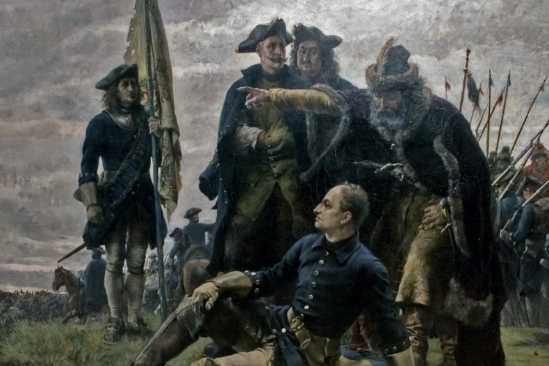 Fem personer i uniform, varav en sitter på marken och tittar mot fjärran, ser ut att diskutera medan en armé av soldater till häst skymtar bakom dem på fältet.