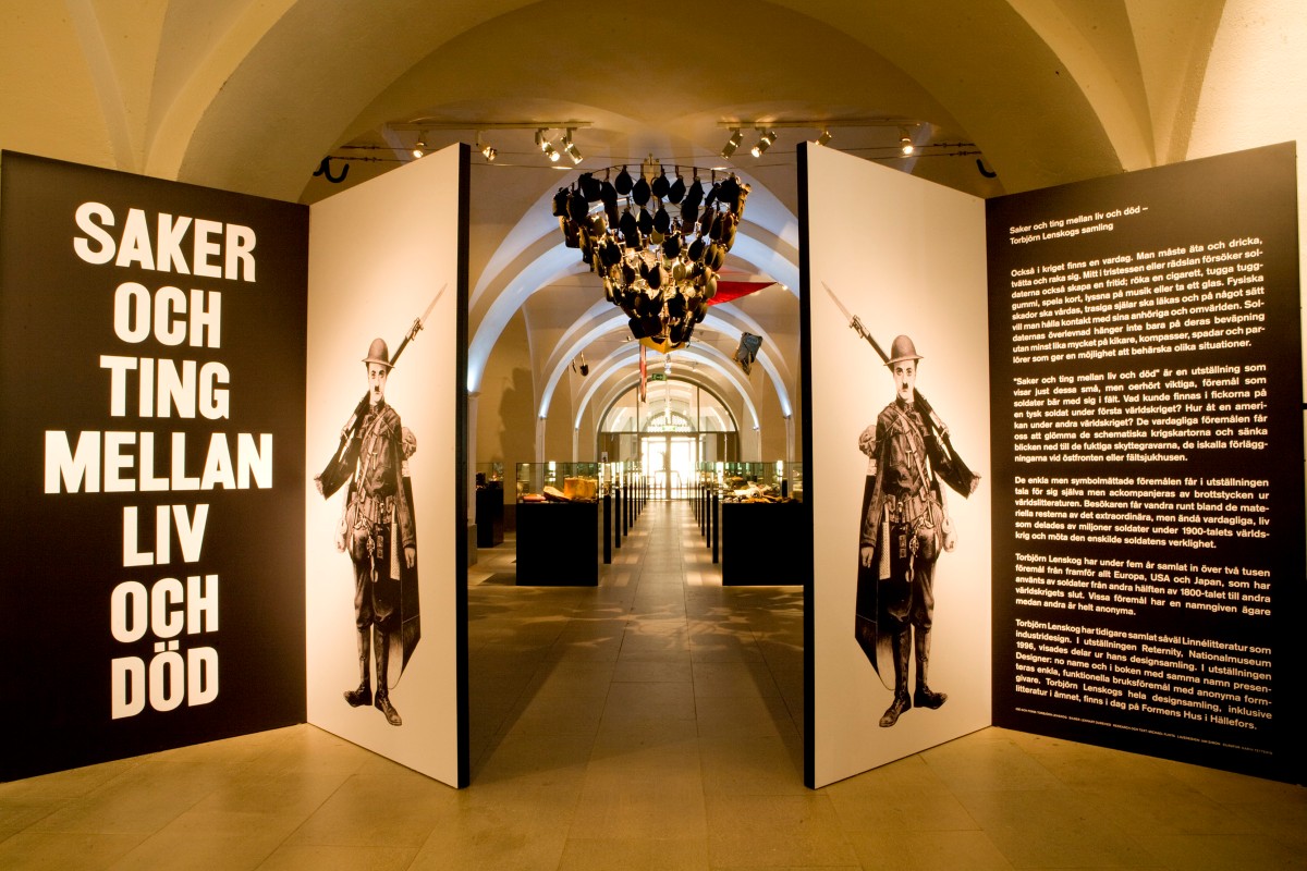 "Saker och ting mellan liv och död" står vid entrén till en utställning. Innanför de öppna dörrarna som pryds av en varsin soldatillustration skymtas glasmontrar med utställningsföremål.