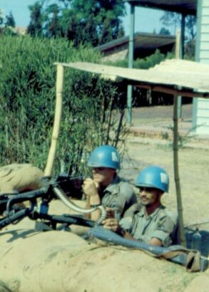 Två soldater med blå hjälmar tittar upp bakom en mur av bruna sandsäckar. Bredvid dem är det buskage och bakom dem ett trähus.