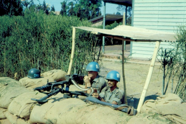 Två soldater med blå hjälmar tittar upp bakom en mur av bruna sandsäckar. Bredvid dem är det buskage och bakom dem ett trähus.