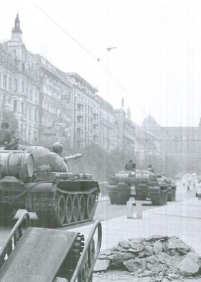 Två stridsvagnar kör genom en stad, en person i rock och med portfölj står bredvid och tittar på.