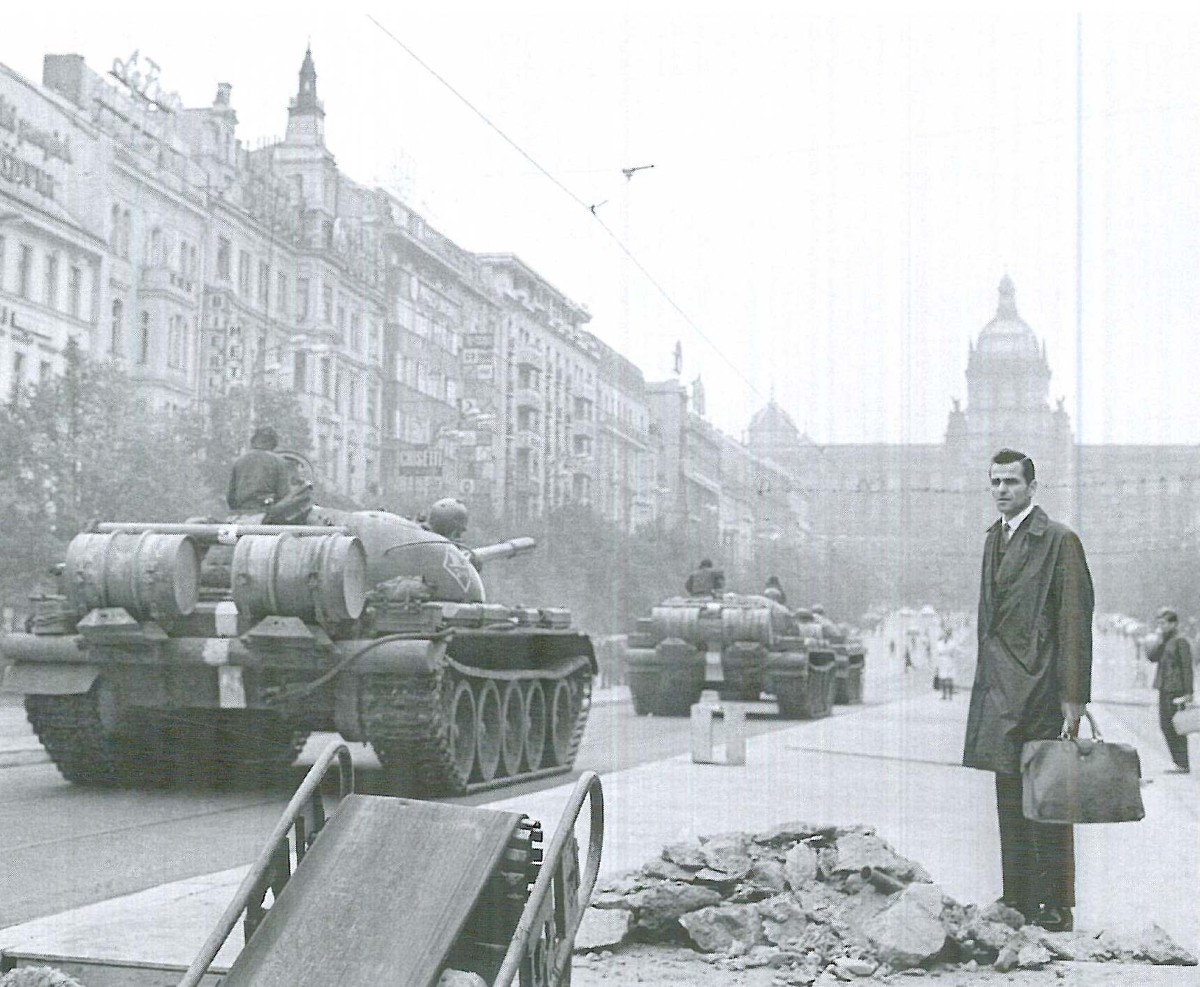Två stridsvagnar kör genom en stad, en person i rock och med portfölj står bredvid och tittar på.