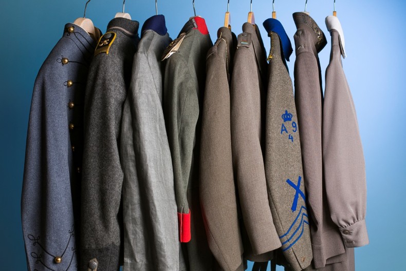 Nio uniformsjackor hänger på galgar på rad. Små detaljer skiljer dem åt såsom olika grå nyanser, ett rött slag, blå revärer.