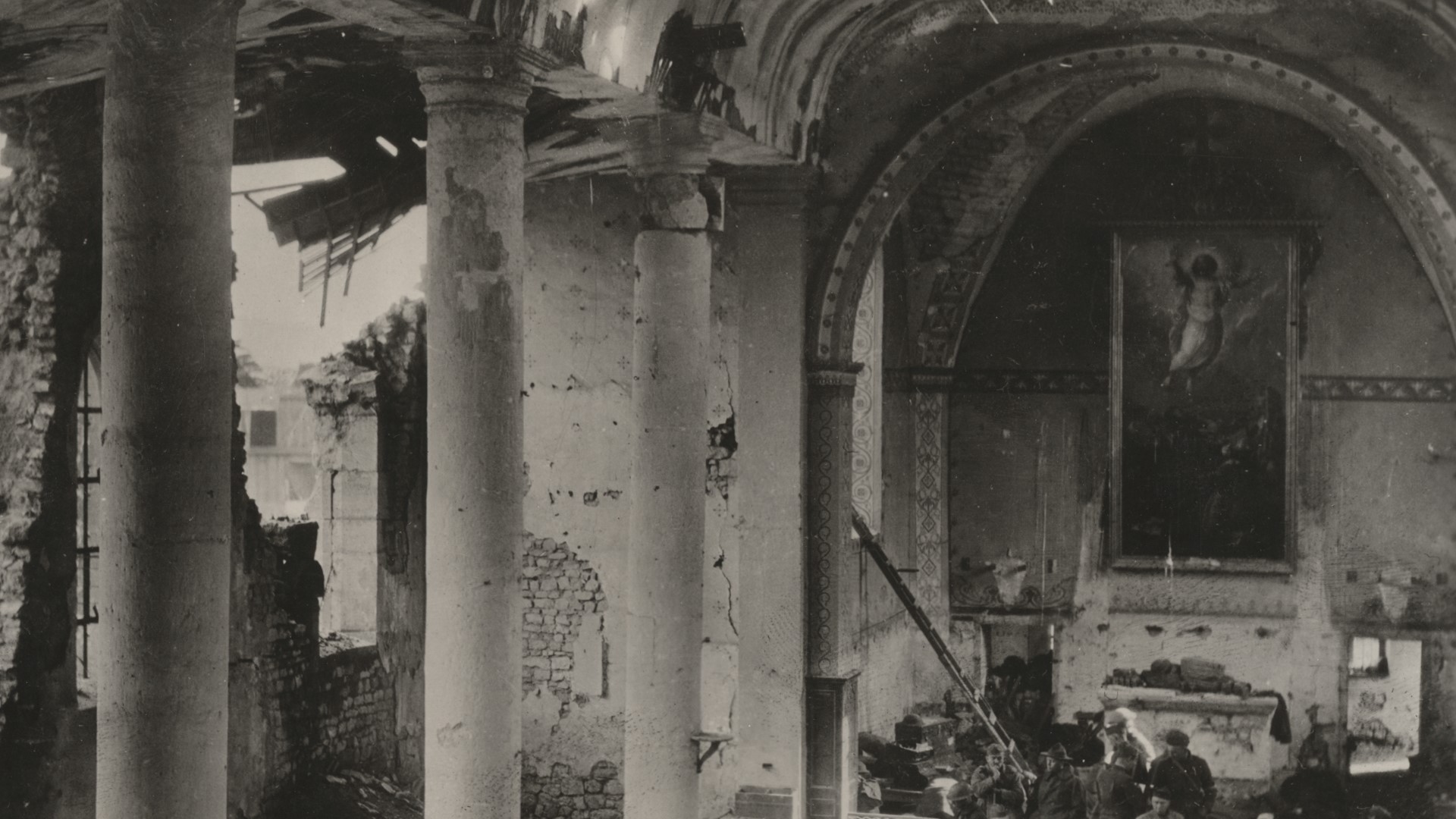 En pampig byggnad med pelare och valv har sargats av bomber. Det var troligtvis en kyrka då det hänger en stor tavla med religiöst motiv på en vägg.