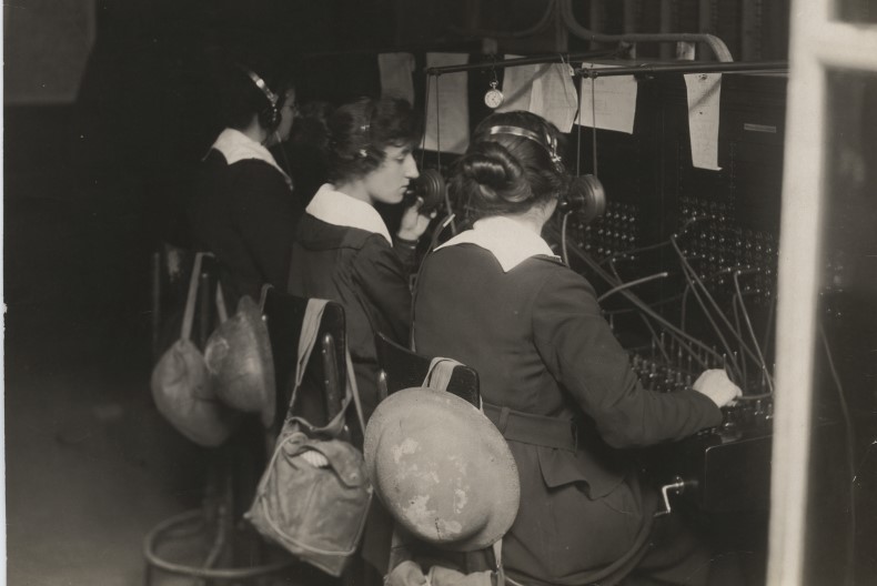 Tre telefonister med hörlurar på sitter med ryggarna mot fotografen. Framför dem växlingsbord med sladdar och knappar. På stolarna har de hängt sina väskor.