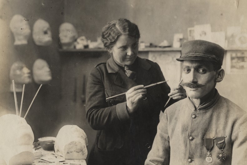 En del av ett rum, på väggarna sitter gipsansikten/huvuden och på en hylla finns ytterligare något huvud. En person i uniform och utmärkelser på bröstet sitter ner och verkar fungera som modell för personen som står upp framför ett bord med ytterliga gipshuvuden.