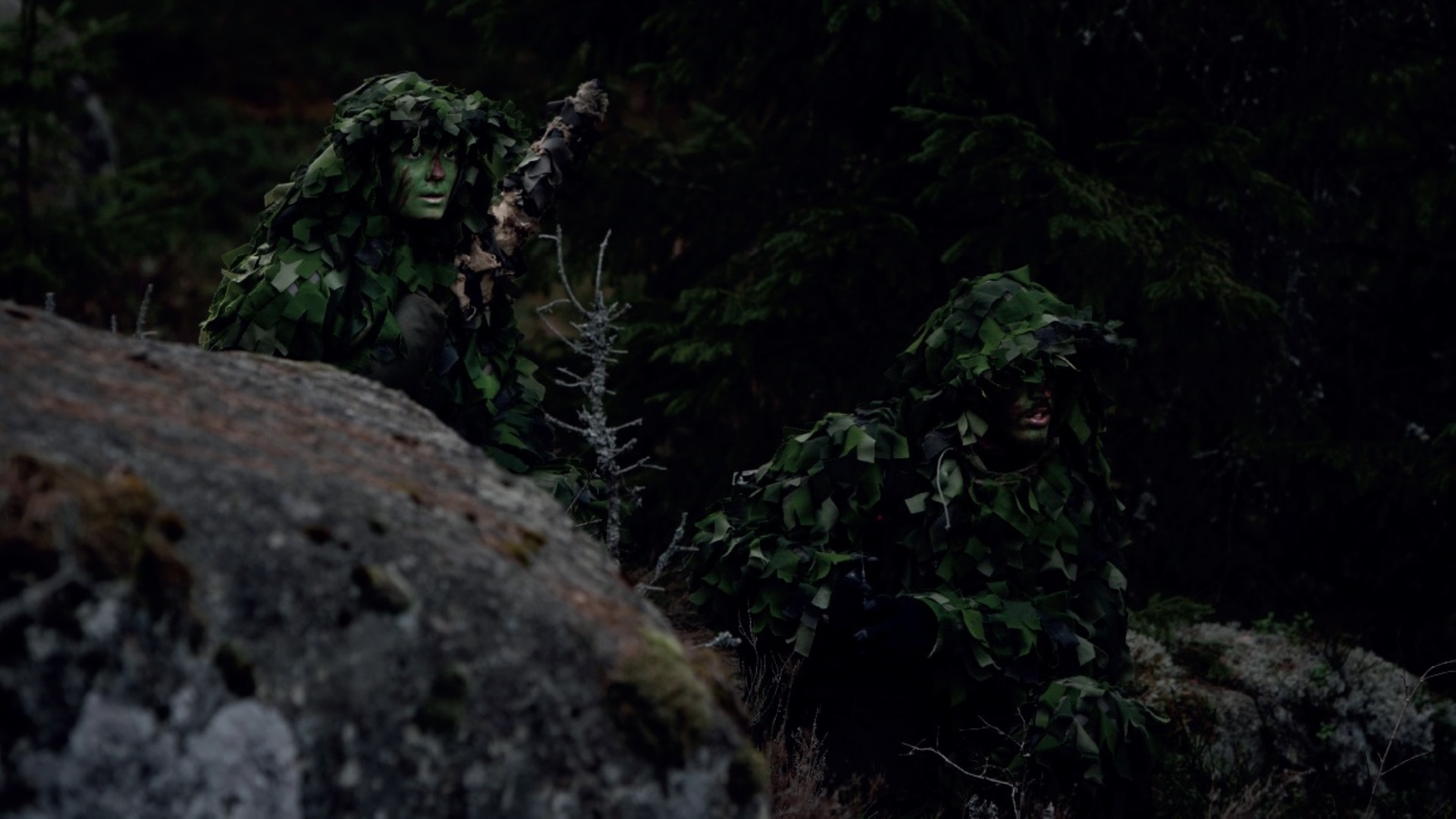 Kamouflageklädda och grönmålade i ansiktena gömmer sig några militärer i skogen och spejar ut ur bild.