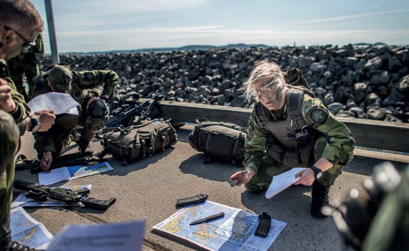 Tre kamouflageklädda militärer sitter på huk och studerar kartor. Omkring dem ligger deras packning samt vapen. Bakom dem en massa större stenar och havet som skymtas.