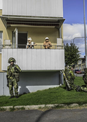 Ett äldre par står på en balkong i ett hyreshus, nedanför dem väntar fyra kamouflageklädda personer med vapen.