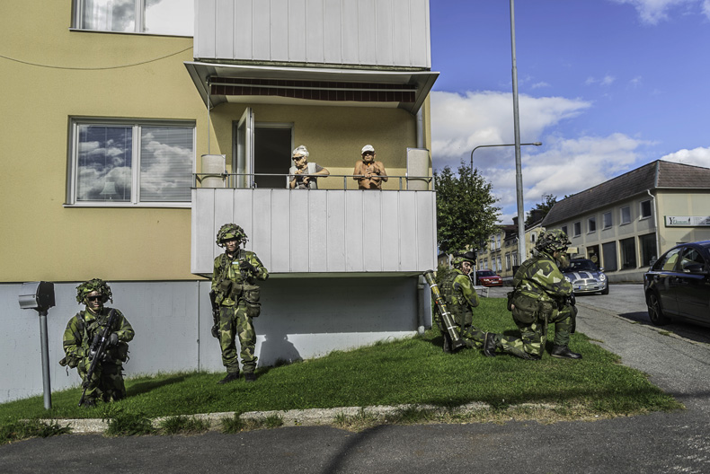 Ett äldre par står på en balkong i ett hyreshus, nedanför dem väntar fyra kamouflageklädda personer med vapen.