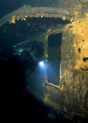 Ett del av ett vrak under vatten och en dykare som lyser med en lampa in i en öppning.