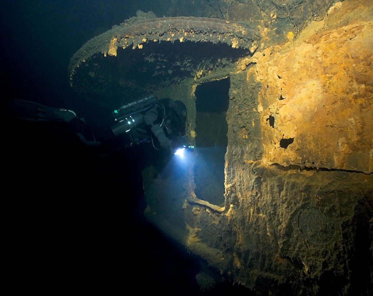 Ett del av ett vrak under vatten och en dykare som lyser med en lampa in i en öppning.
