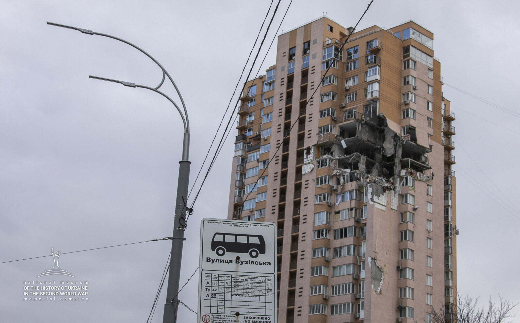 Ett höghus som har ett stort hål i ena sidan där det troligtvis har farit in en bomb. I förgrunden en busshållplatsskylt.