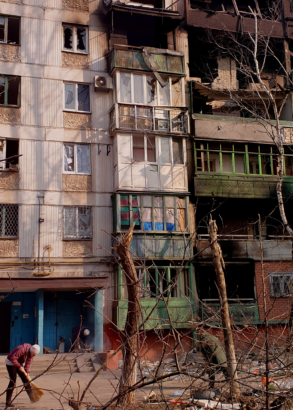 Två människor sopar gården framför ett sönderbombat hus. Skräp på marken, sönderslitna träd.